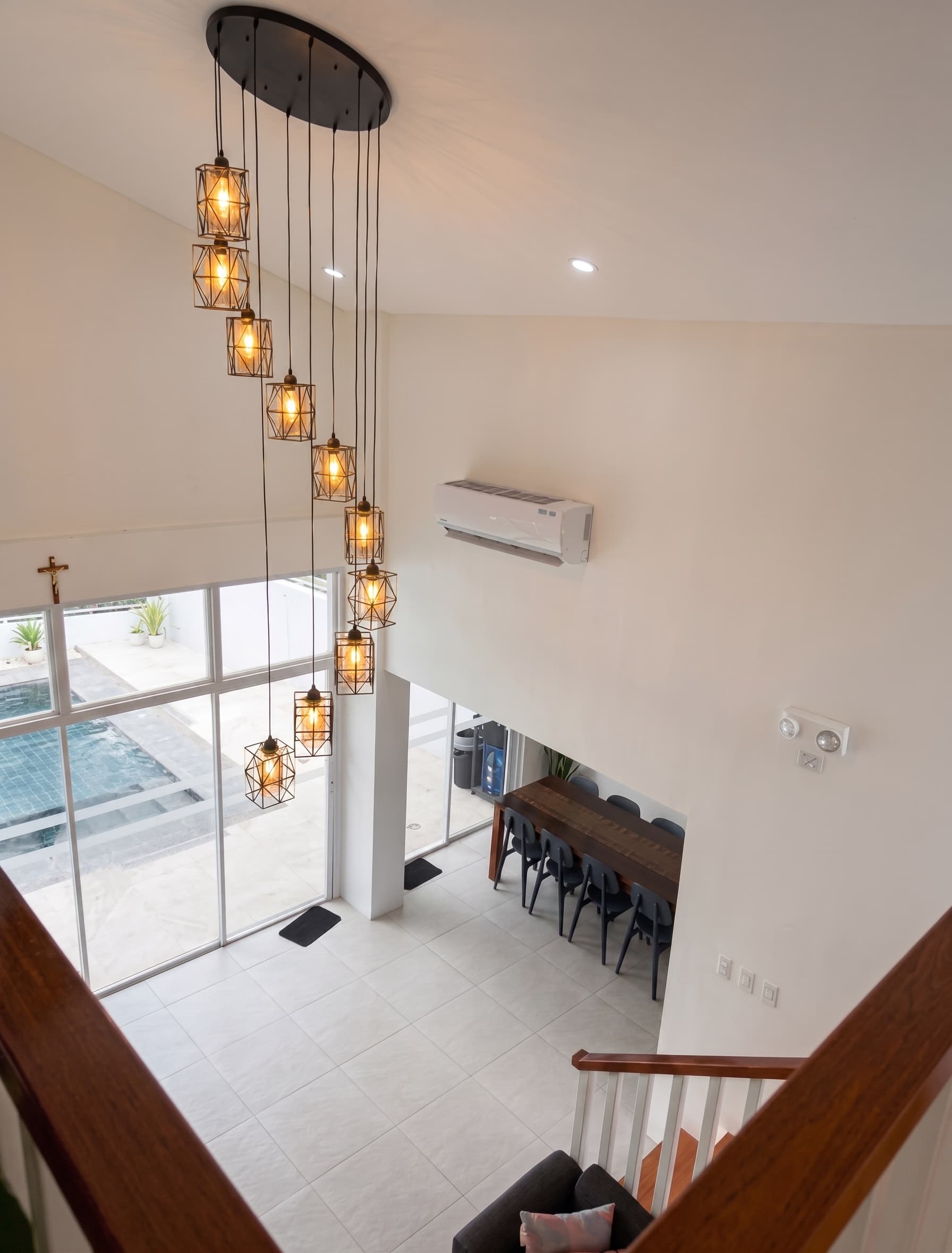 Looking down from the stairs at the double-height living room, its cluster of pendant lights, and the pool through the glass doors
